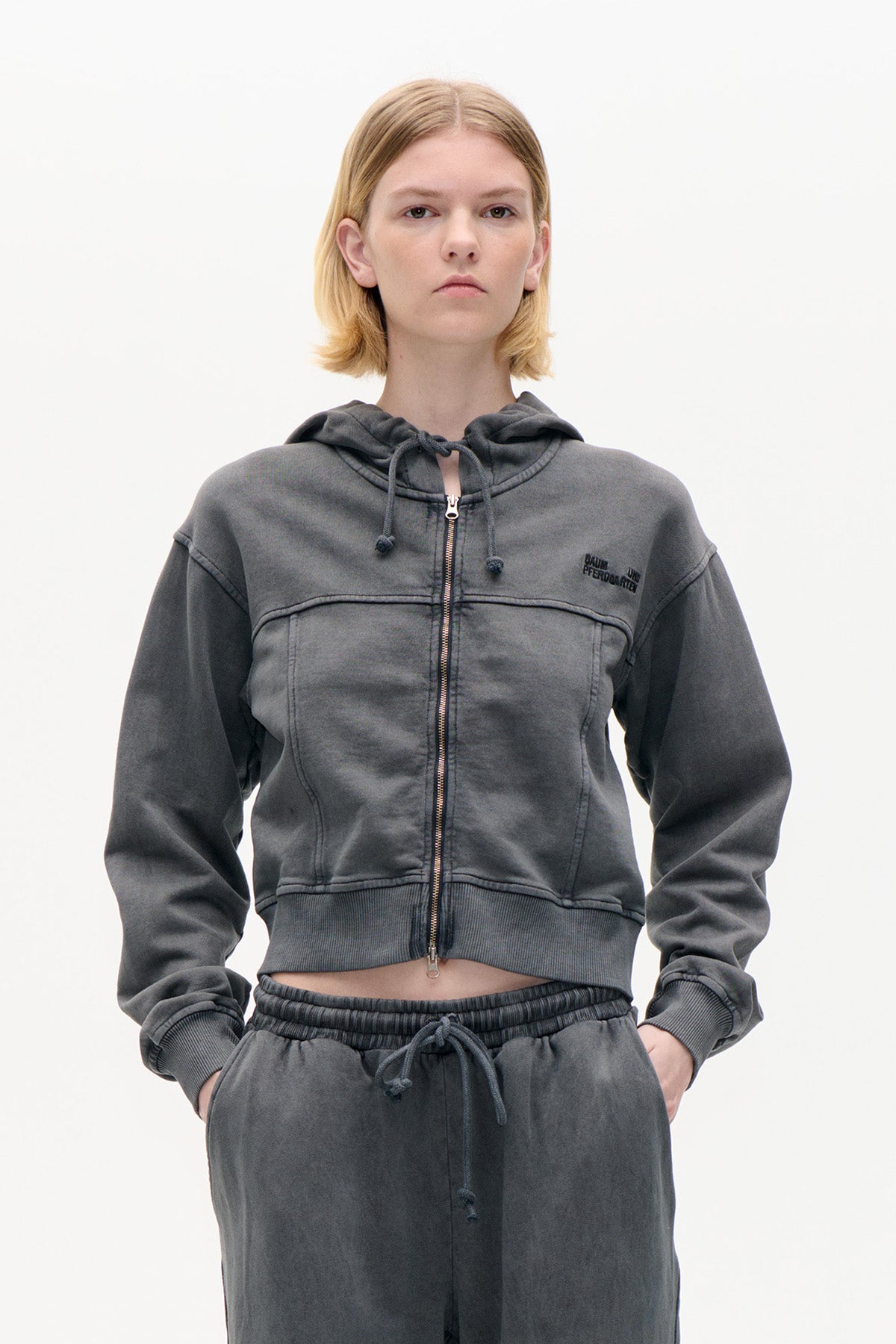 Front view of a model wearing a charcoal grey zip-up hoodie with drawstring hood and chest logo, paired with matching drawstring trousers.