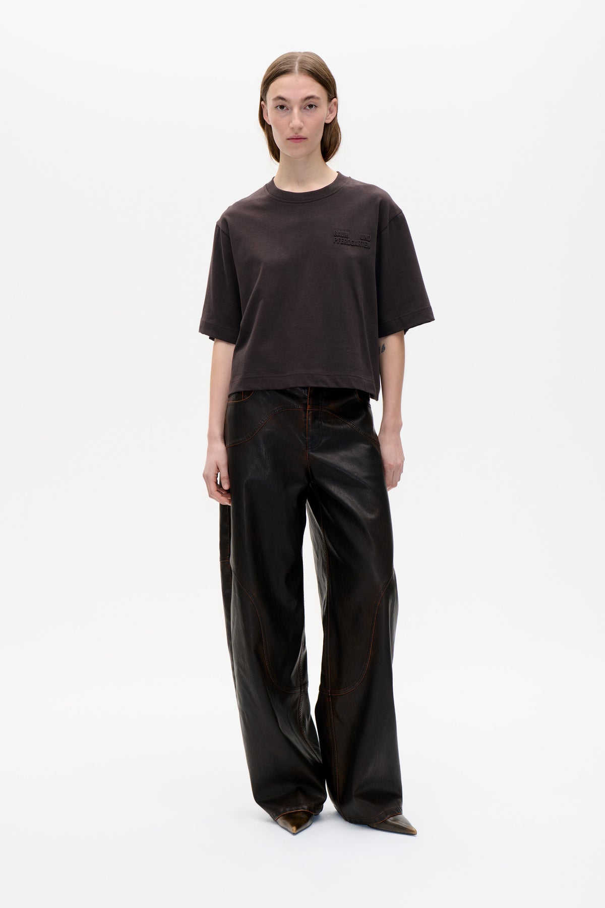 Model trägt ein dunkelbraunes, oversized T-Shirt mit kurzen Ärmeln und tonaler Brustschrift sowie eine schwarze, weit geschnittene Hose mit geschwungener Kontrastnaht.