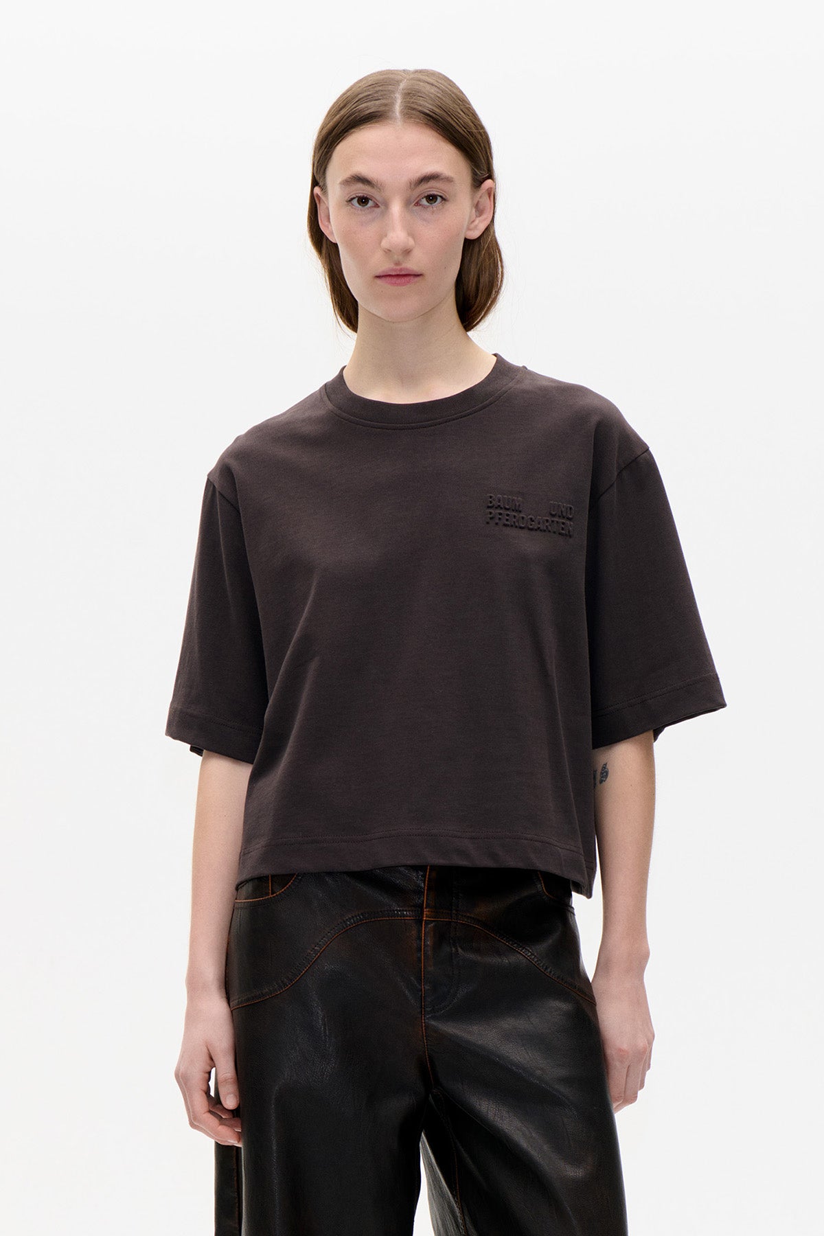 Model trägt ein dunkelbraunes, oversized, kurzärmeliges T-Shirt mit tonalem Brustlogo und eine schwarze Hose in Lederoptik. Frontalansicht.