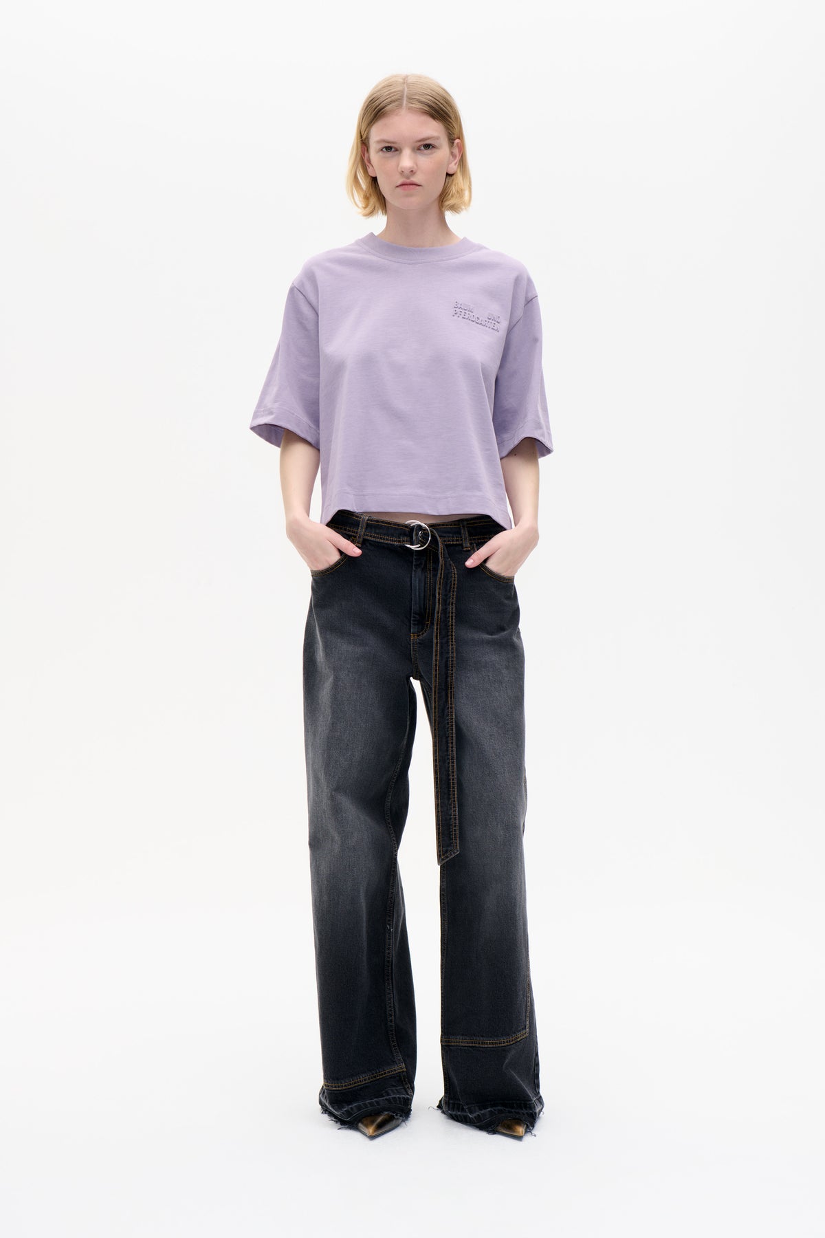 Model trägt ein fliederfarbenes Oversized-Kurzarm-T-Shirt mit erhabenem Schriftzug auf der Brust und anthrazitfarbene Jeans mit weitem Bein und langem Gürtelriemen.