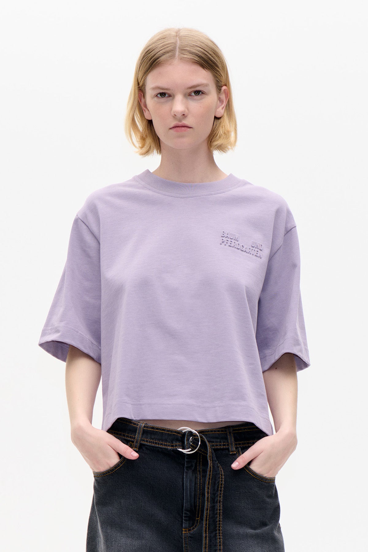 Vorderansicht eines fliederfarbenen T-Shirts mit kurzen Ärmeln, Rundhalsausschnitt und tonalem, erhabenem Brustschriftzug, kombiniert mit dunklen Jeans.
