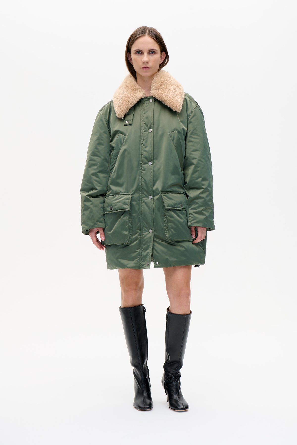 Front view of a model wearing a long sage green coat with a cream plush collar, snap-front fastening and large flap pockets.