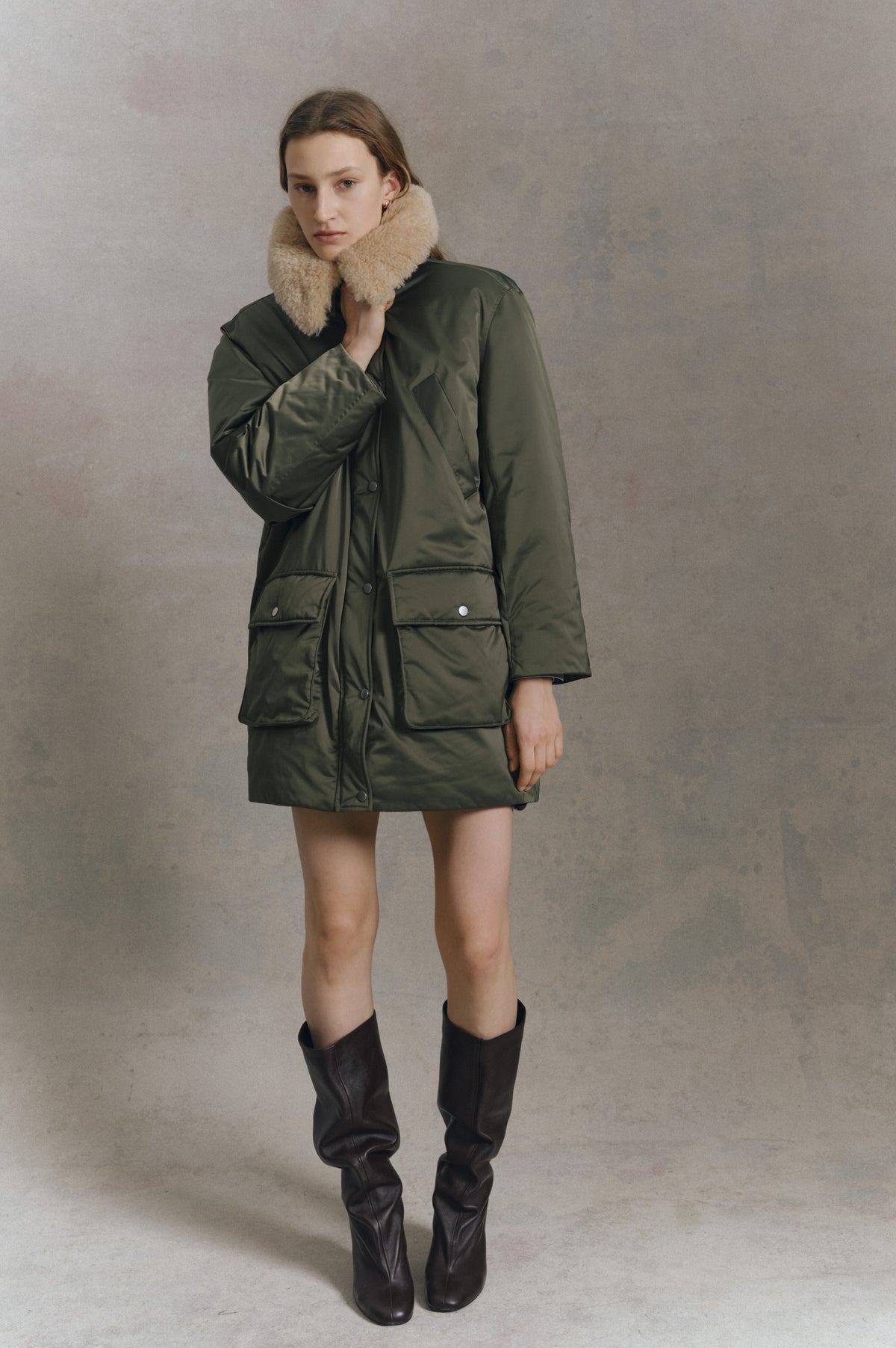 Model wearing a dark olive knee-length coat with snap closures, large patch pockets and a plush beige collar, styled with tall dark boots.