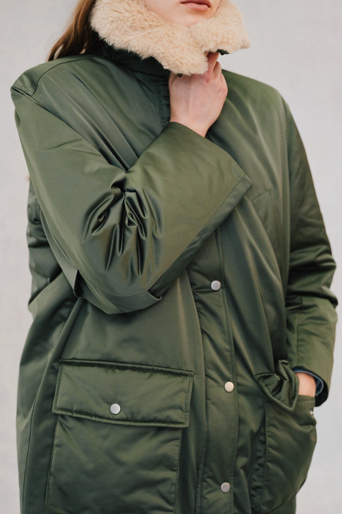 Close-up campaign portrait of a model wearing an olive green utility jacket with snap-button flap pockets and a thick cream fuzzy scarf.