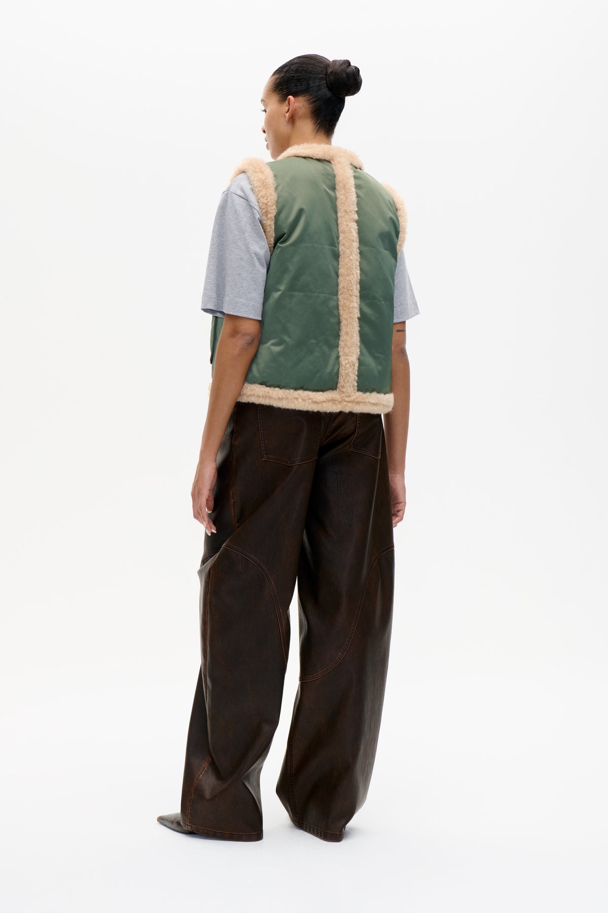Model viewed from behind wearing a sage green sleeveless vest with cream fluffy trim, a grey T-shirt and dark brown wide-leg trousers.