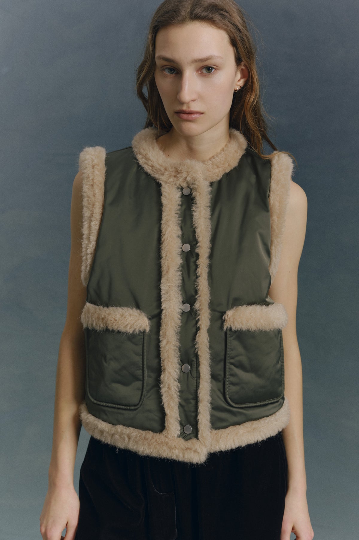 Close-up campaign portrait of a model wearing a dark olive padded vest with cream fluffy trim, snap buttons and front patch pockets.