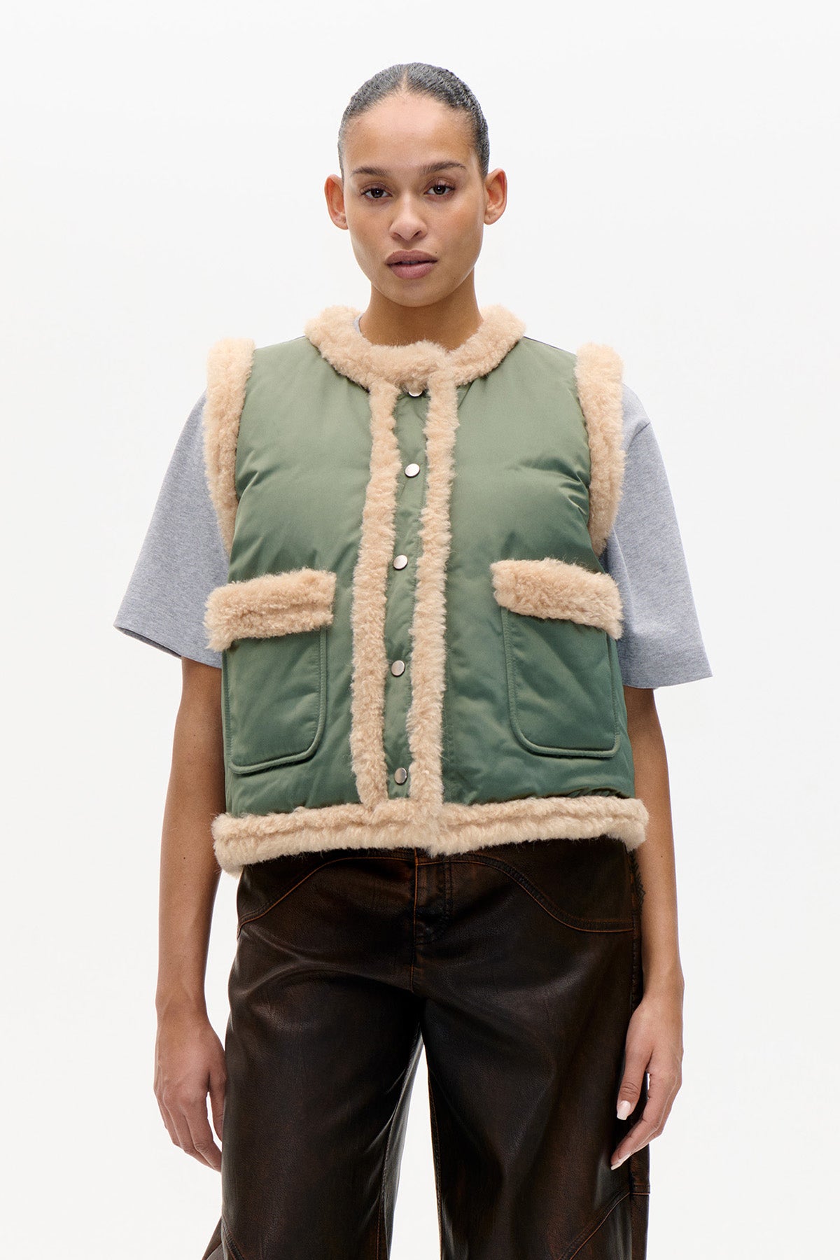 Front view of a sage green sleeveless vest with cream plush trim, snap buttons and patch pockets, layered over a grey T-shirt.