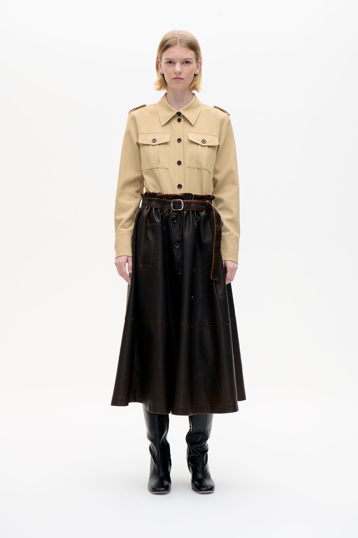 Front-facing model in a beige button-front shirt with pointed collar and flap pockets, paired with a glossy black belted skirt and knee-high boots.