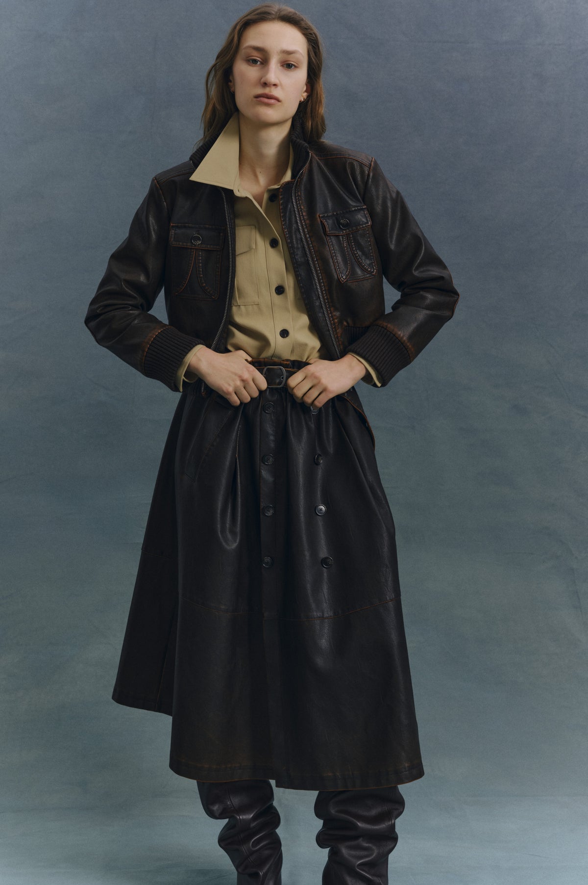 Campaign portrait of a model wearing a dark brown leather-look jacket with contrast stitching and ribbed cuffs over a beige collared shirt.