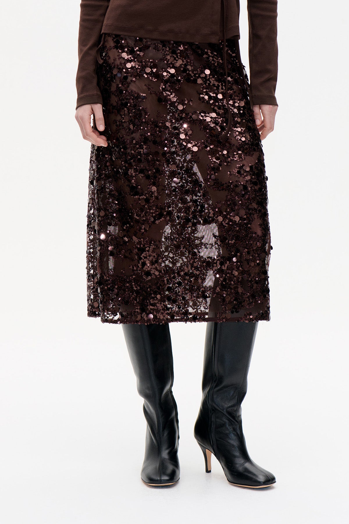 Close-up of a dark brown knee-length skirt covered in scattered sequins, styled with a brown top and black knee-high boots.