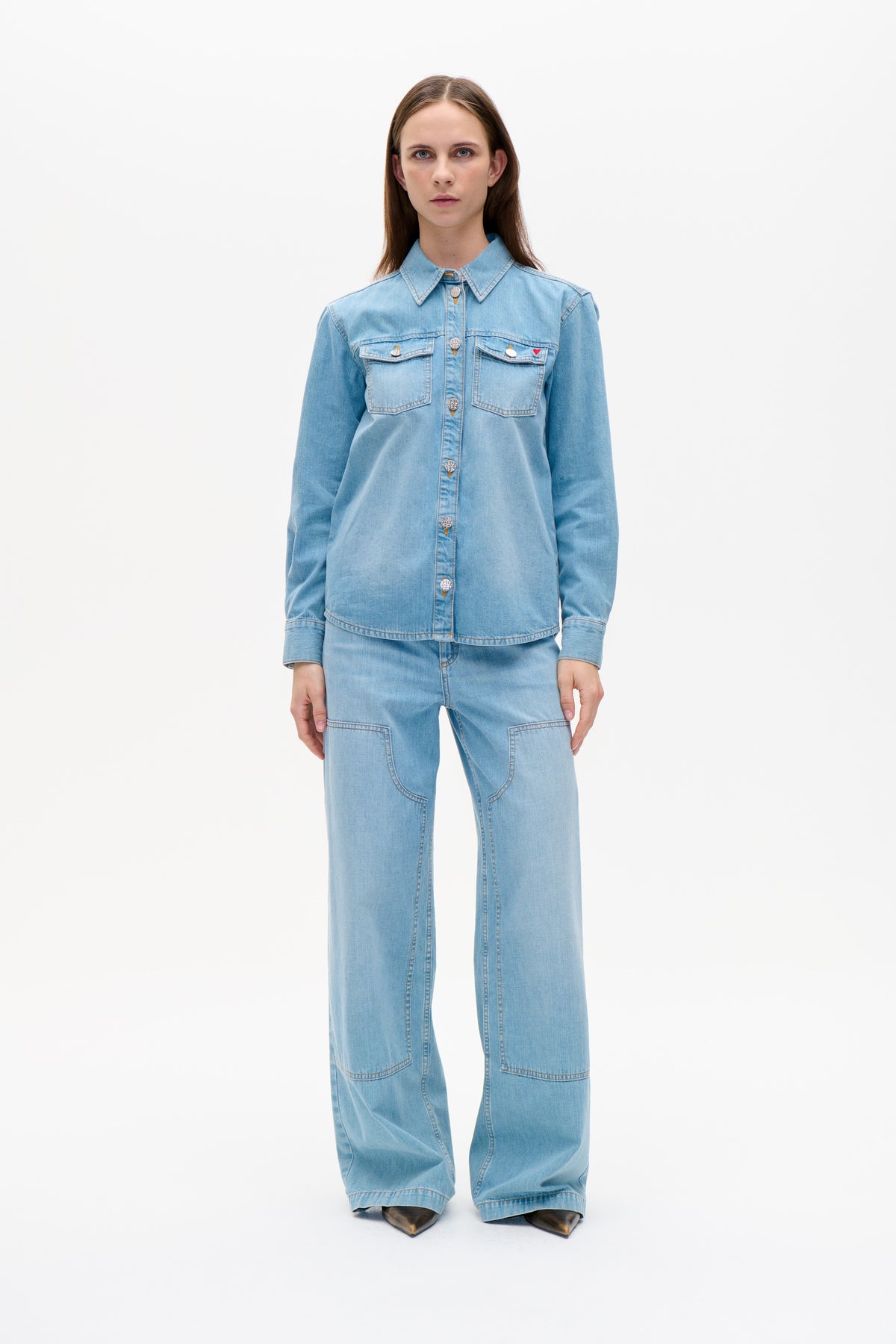 Model wearing a light blue denim shirt with pointed collar, chest pockets and decorative pins, paired with wide-leg jeans.