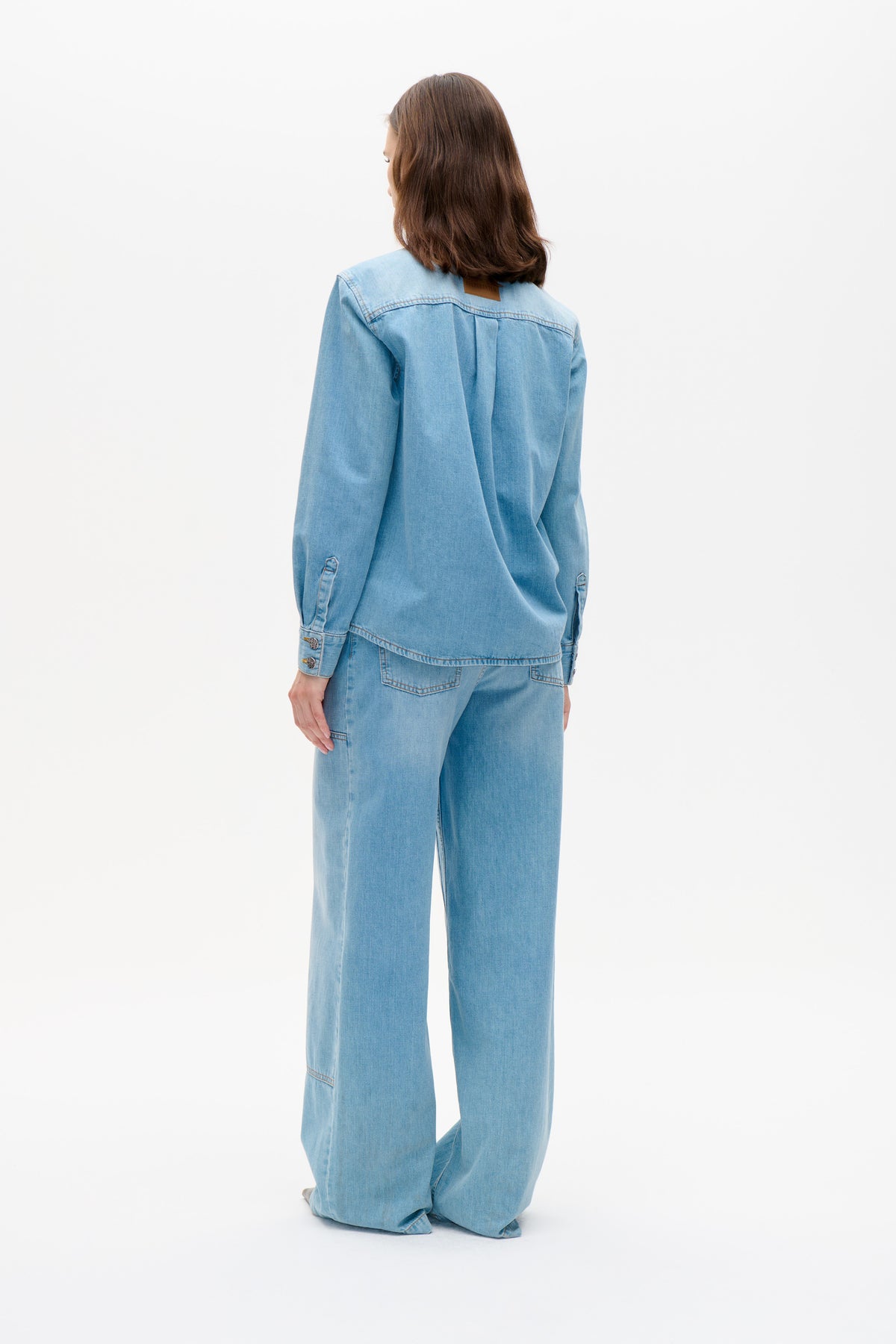 Back view of a model wearing a light blue denim jacket with a yoke seam and center pleat, paired with wide-leg jeans.