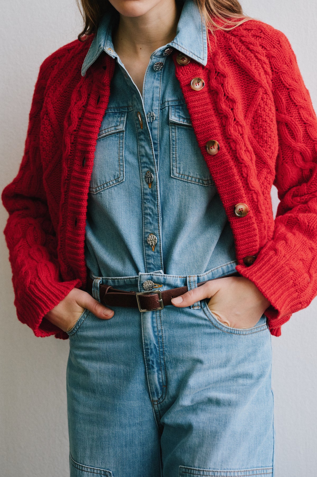 Cropped campaign portrait showing a model in a bright red textured knit cardigan over a light blue denim button-front outfit with a belt.