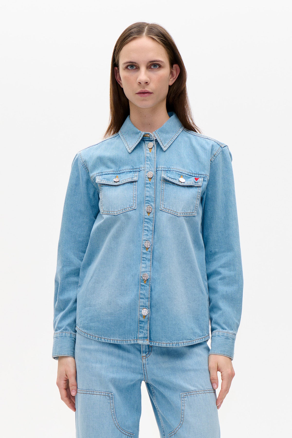Front view of a light blue denim button-up shirt with pointed collar, two flap chest pockets and decorative buttons, worn with matching jeans.