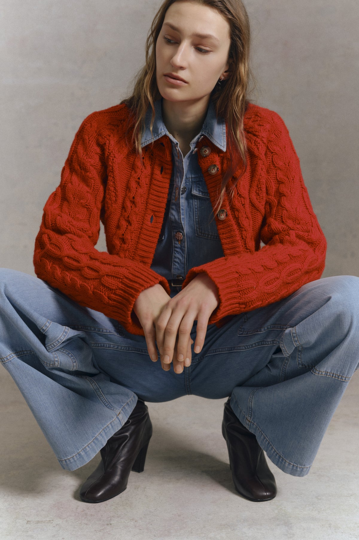 Campagneportret van een zittend model dat een felrode, kabelgebreide cardigan met knoopsluiting draagt, gecombineerd met een blauwe denimblouse en jeans.