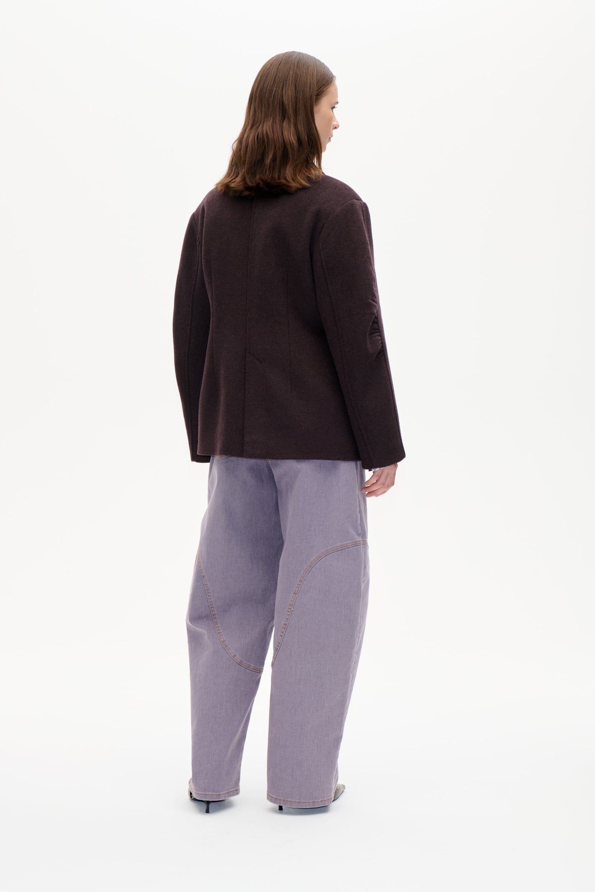 Back view of a model wearing a dark brown tailored jacket with seam detailing and pale lavender wide-leg jeans.