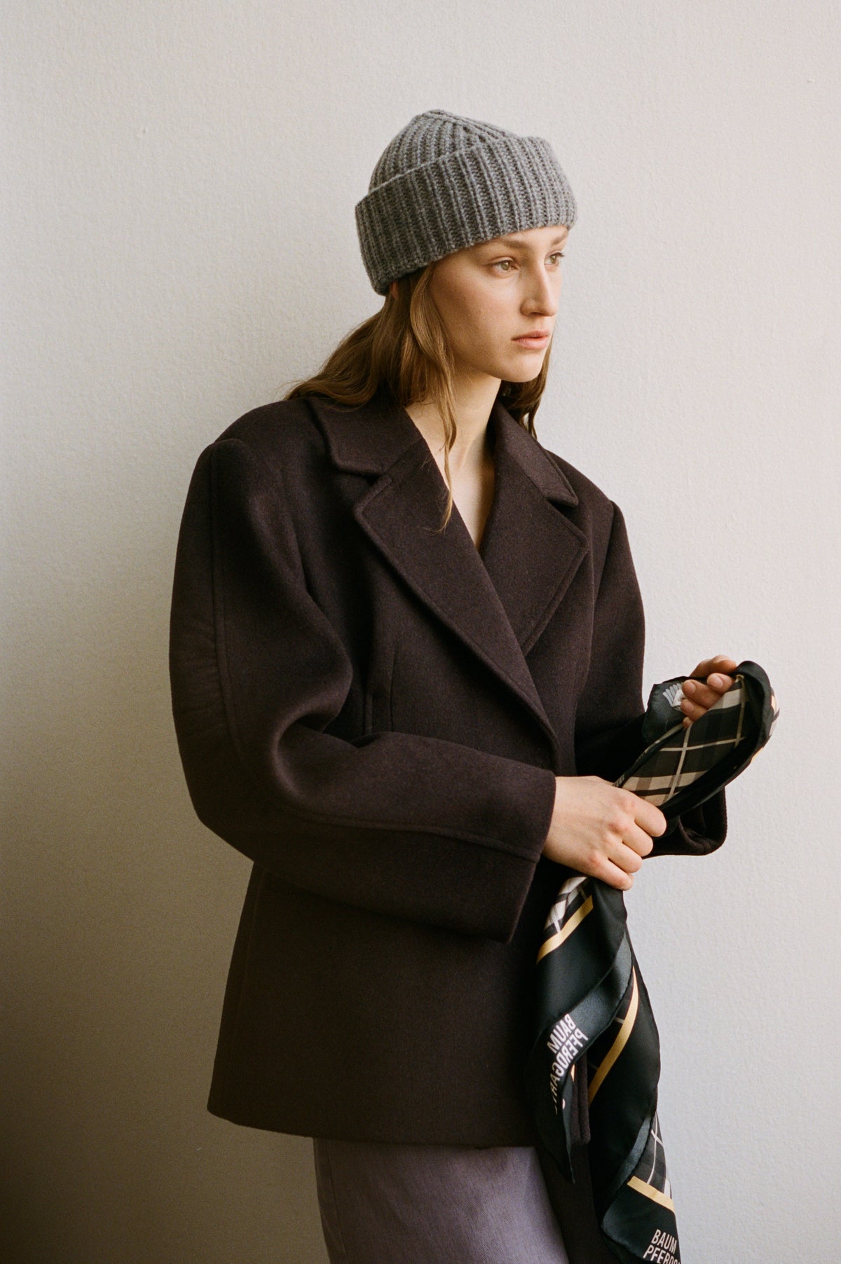 Campaign portrait of a model wearing a grey ribbed knit beanie and dark double-breasted coat, holding a black patterned scarf.
