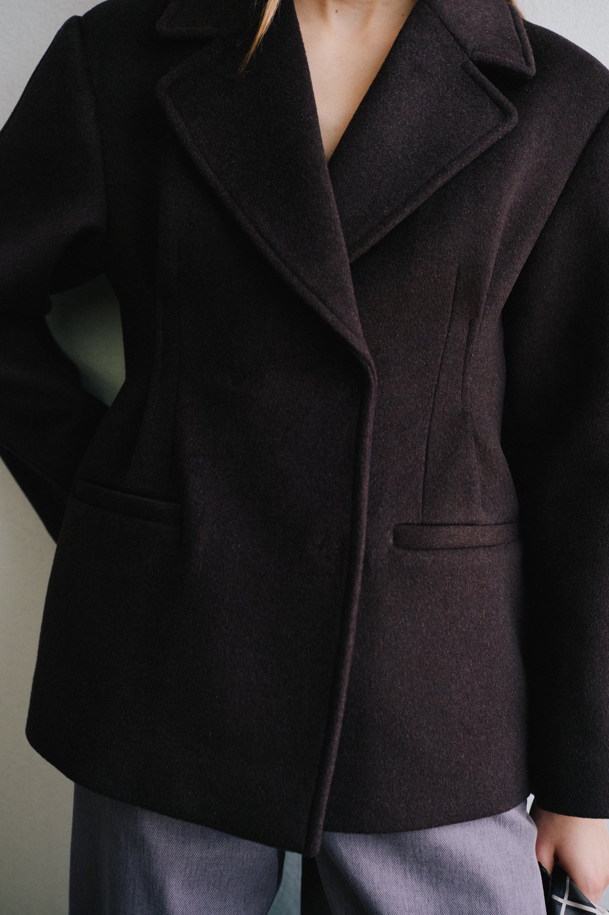 Close-up campaign view of a model wearing a dark charcoal-brown jacket with wide lapels and a flap pocket, paired with grey trousers.