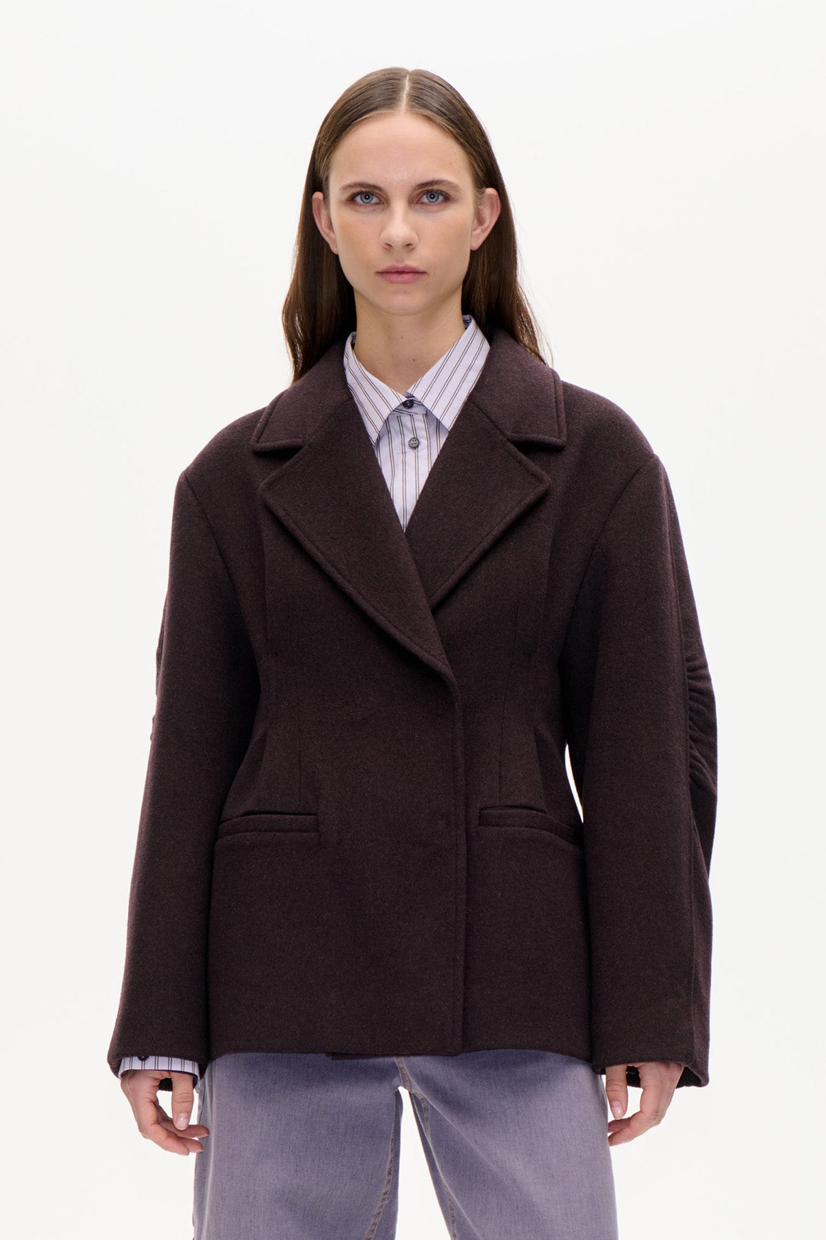 Front view of a dark brown oversized double-breasted coat with wide lapels and flap pockets, layered over a striped shirt and pale jeans.
