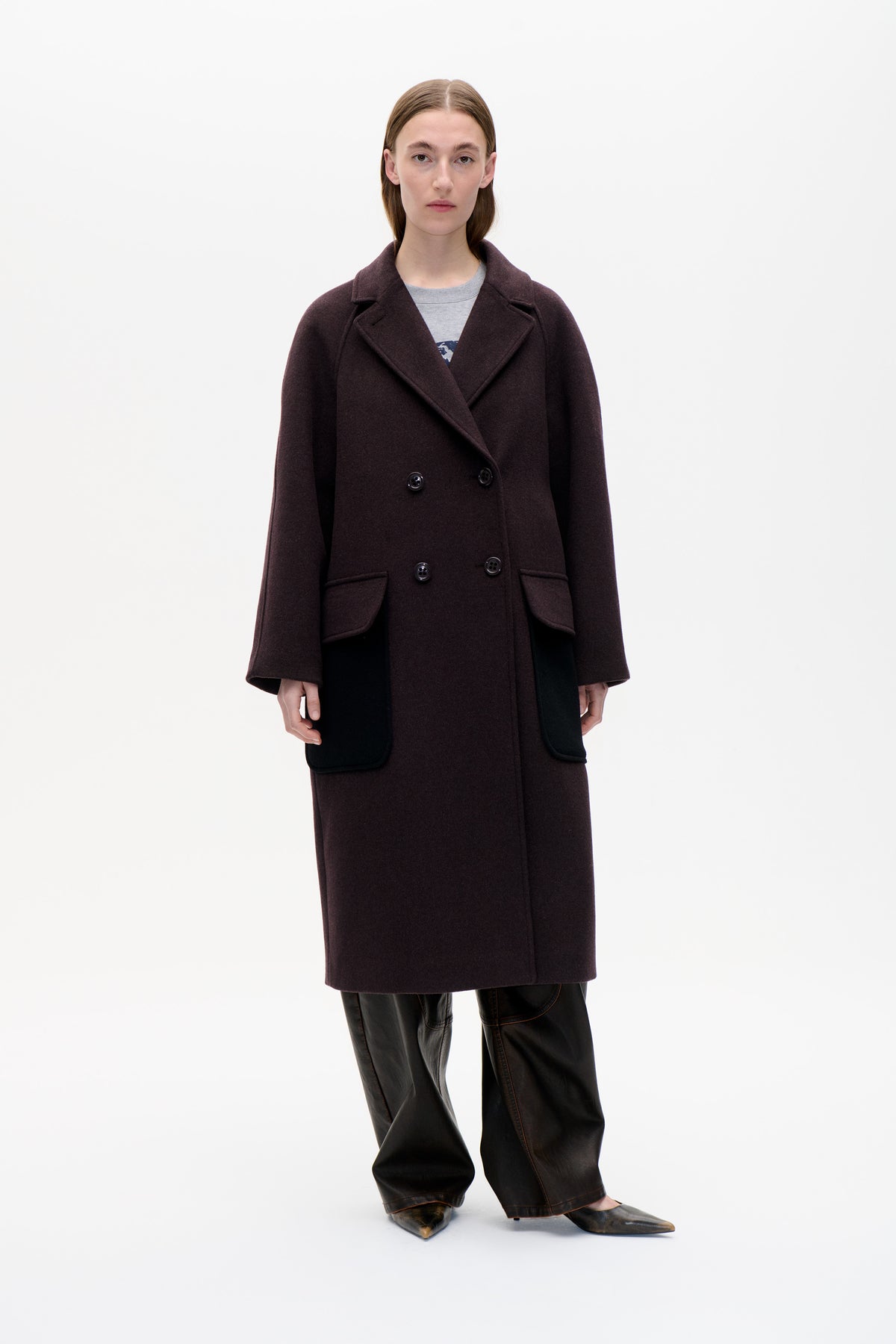 Front view of a model wearing a dark plum double-breasted coat with wide lapels and flap pockets over a grey top.