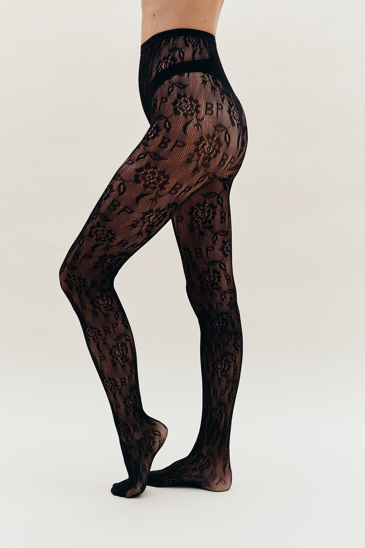 Close view of sheer black tights with an all-over floral lace pattern, shown on crossed legs from waist to feet.