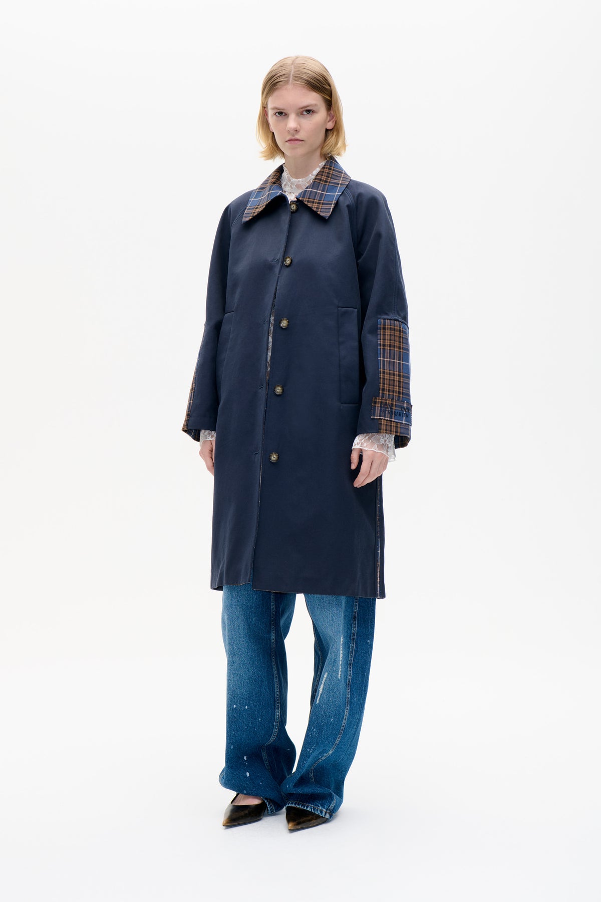 Model wearing a long navy coat with brown and blue plaid collar and cuff panels, layered over a white lace top and blue jeans.