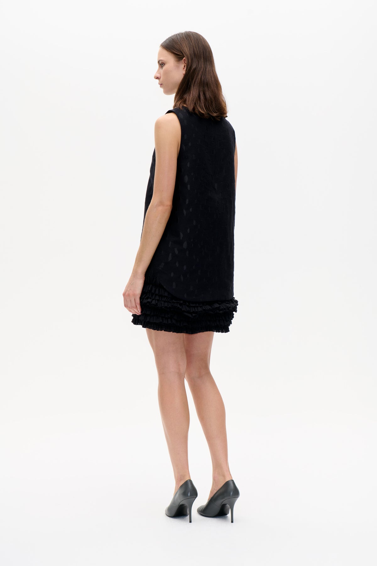 Model in side-back view wearing a black sleeveless mini dress with a subtle tonal pattern and layered ruffle hem.