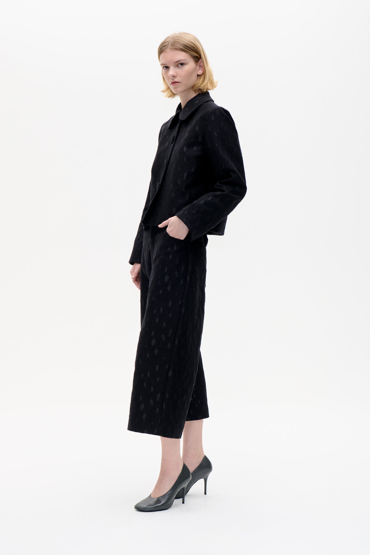 Model wearing a black button-front jacket with a rounded collar and matching knee-length skirt in a subtle tonal spot pattern.