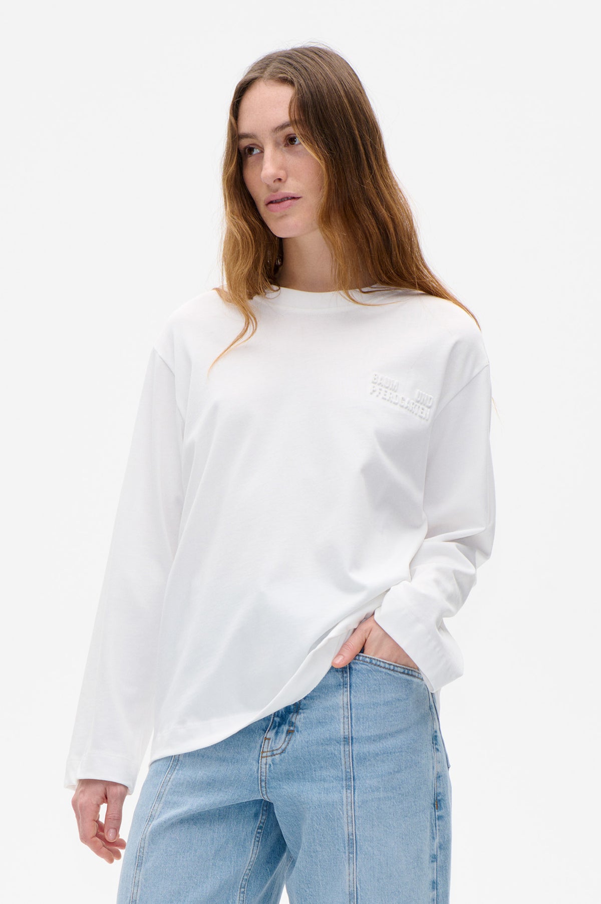 Model wearing a white long-sleeved sweatshirt with raised Baum und Pferdgarten lettering, paired with light-wash blue jeans.