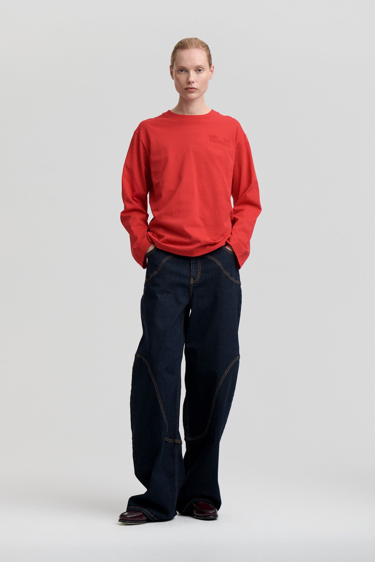 Front view of a model wearing a bright red long-sleeved sweatshirt with subtle chest lettering and dark wide-leg jeans.