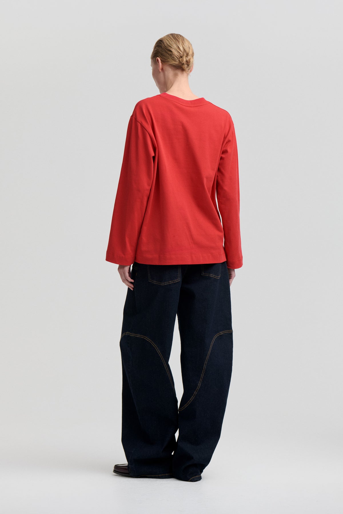 Rückenansicht eines Models, das ein schlichtes rotes Langarm‑Oberteil mit Rundhalsausschnitt und eine dunkle, weit geschnittene Jeans mit Kontrastnähten trägt.
