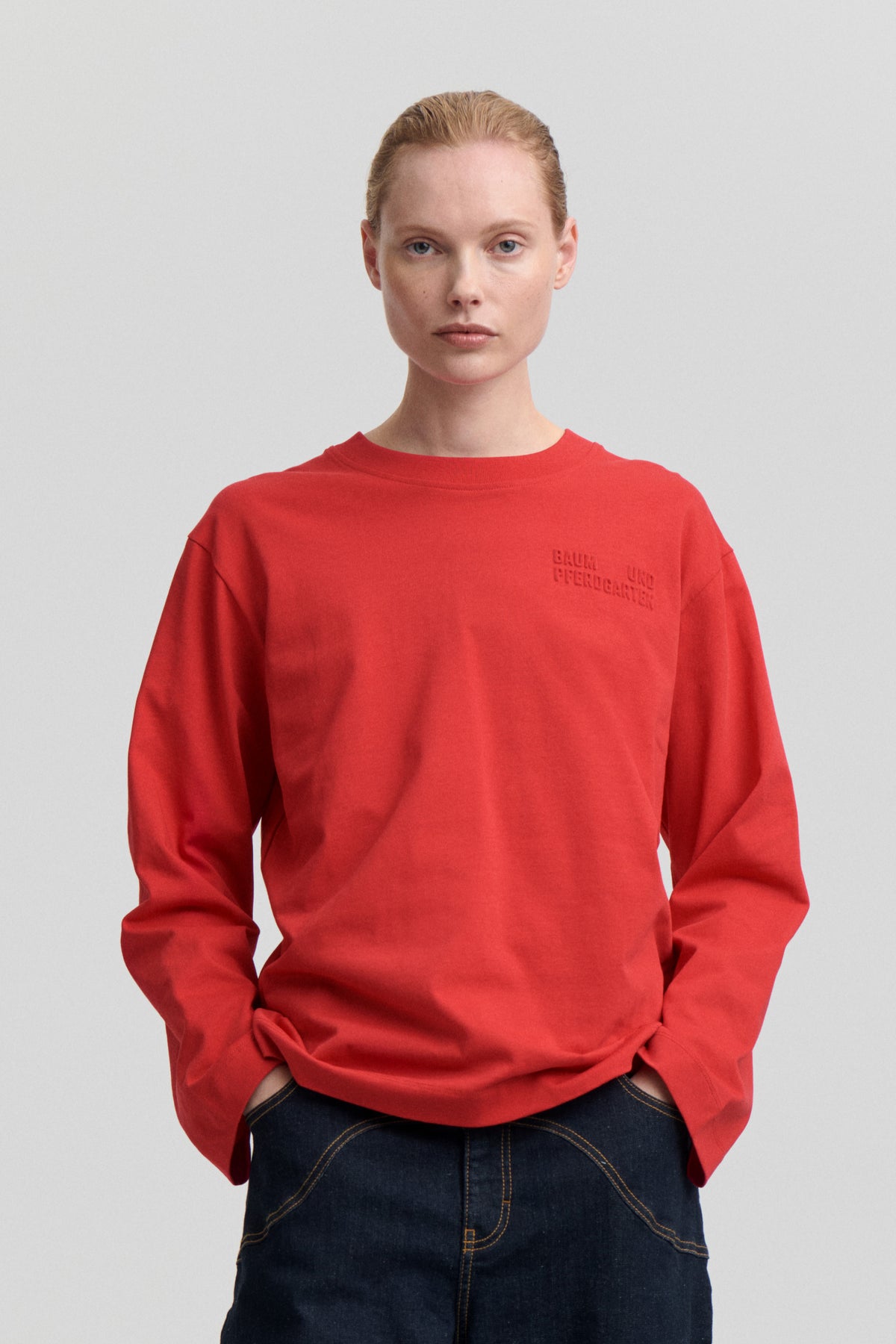 Model wearing a red crew-neck sweatshirt with tonal Baum und Pferdgarten lettering on the chest and dark jeans.