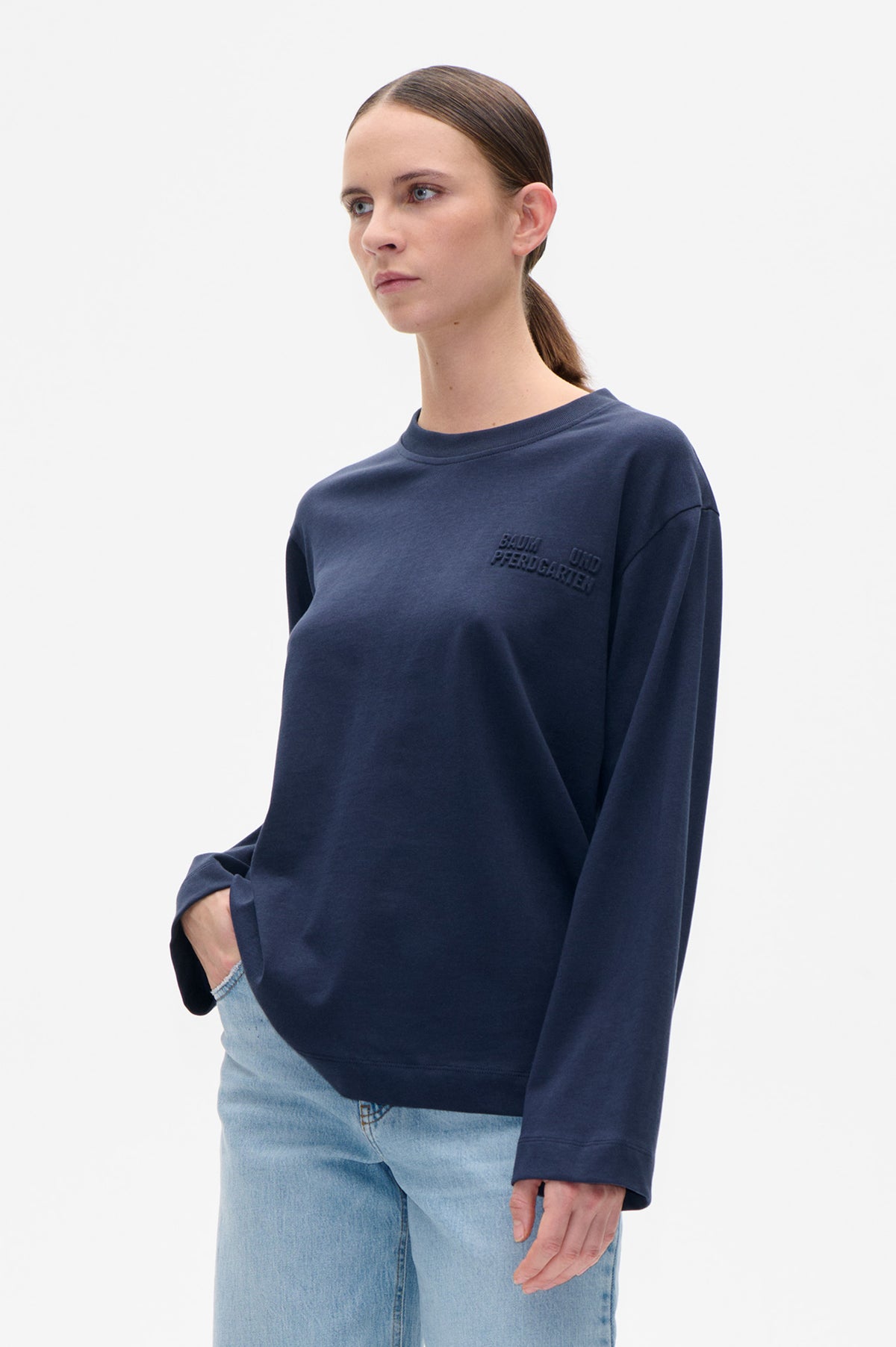 Model trägt ein marineblaues Rundhals-Sweatshirt mit erhabenem Baum und Pferdgarten-Schriftzug und eine hell gewaschene Jeans, von vorne gezeigt.