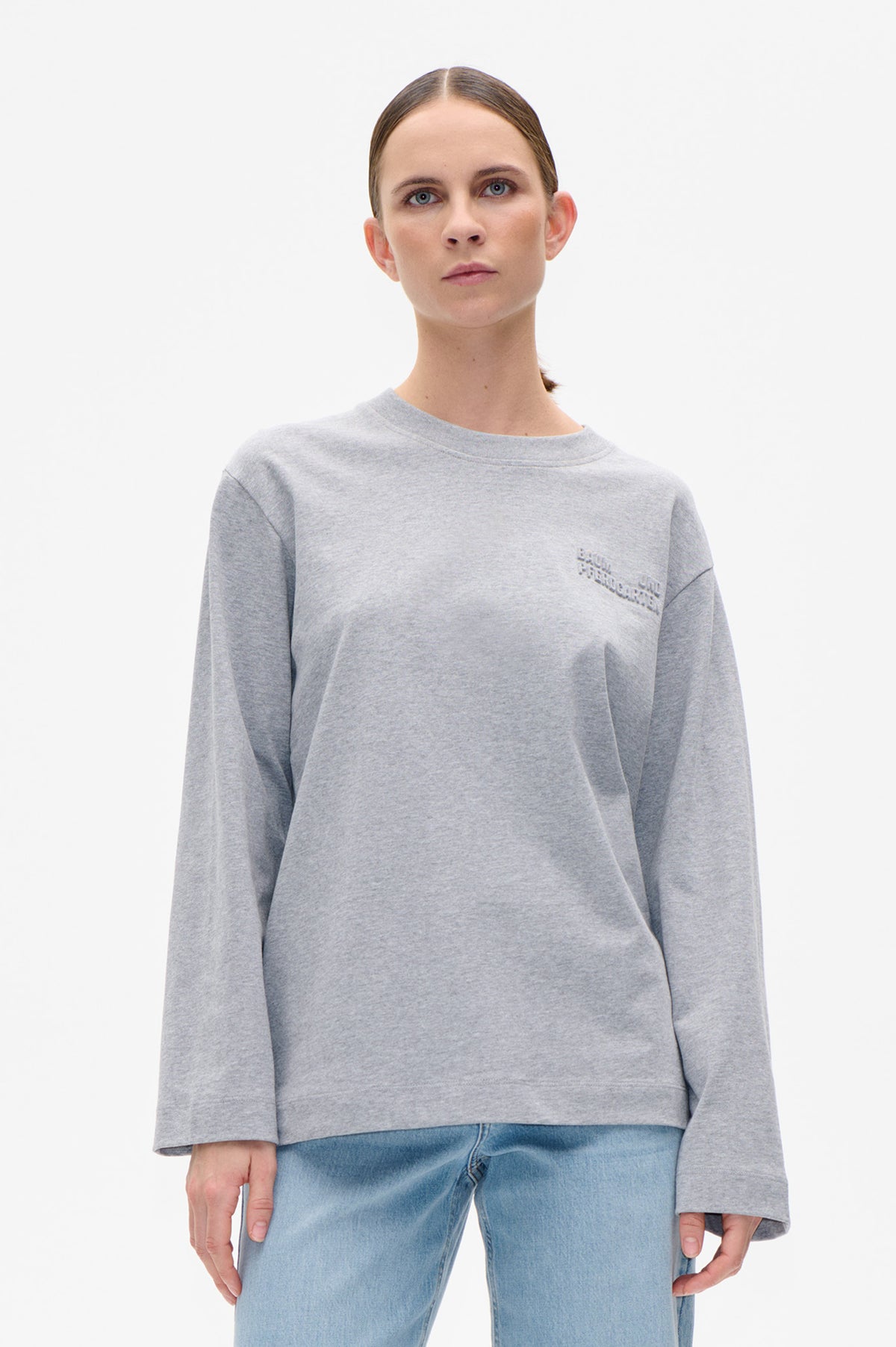 Front view of a model wearing a light grey crew-neck sweatshirt with long sleeves and subtle raised chest lettering, paired with light-wash jeans.