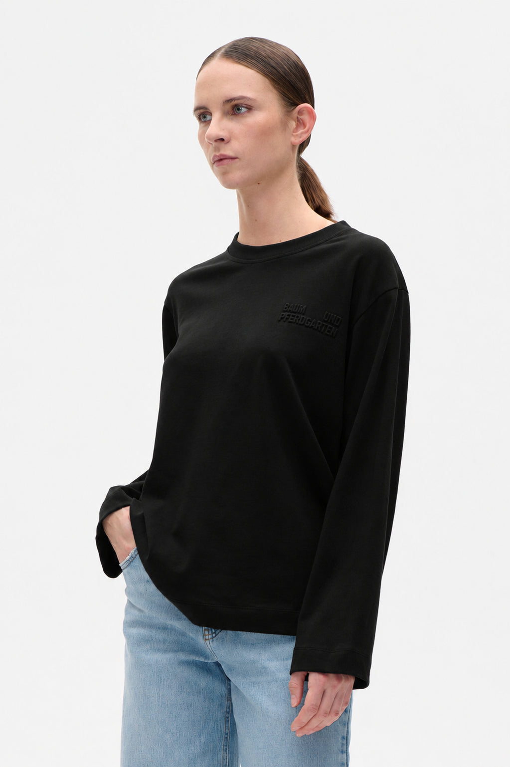 Model trägt ein schwarzes Rundhals-Sweatshirt mit dezenter Brustaufschrift und hellblauen Jeans, von vorne aufgenommen.