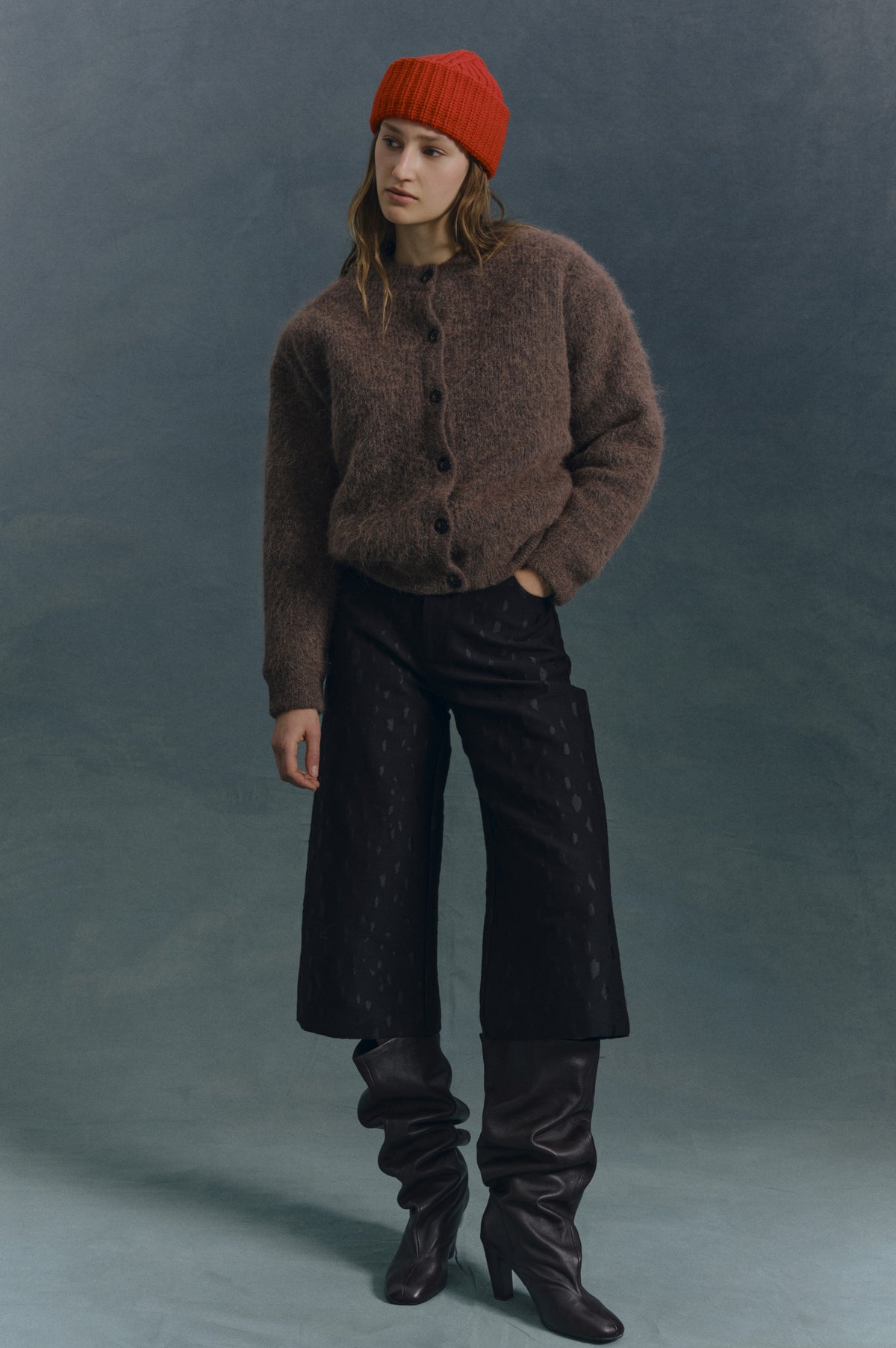 Campaign portrait of a model wearing a bright red rib-knit beanie, brown button-front cardigan, black patterned trousers and tall dark boots.