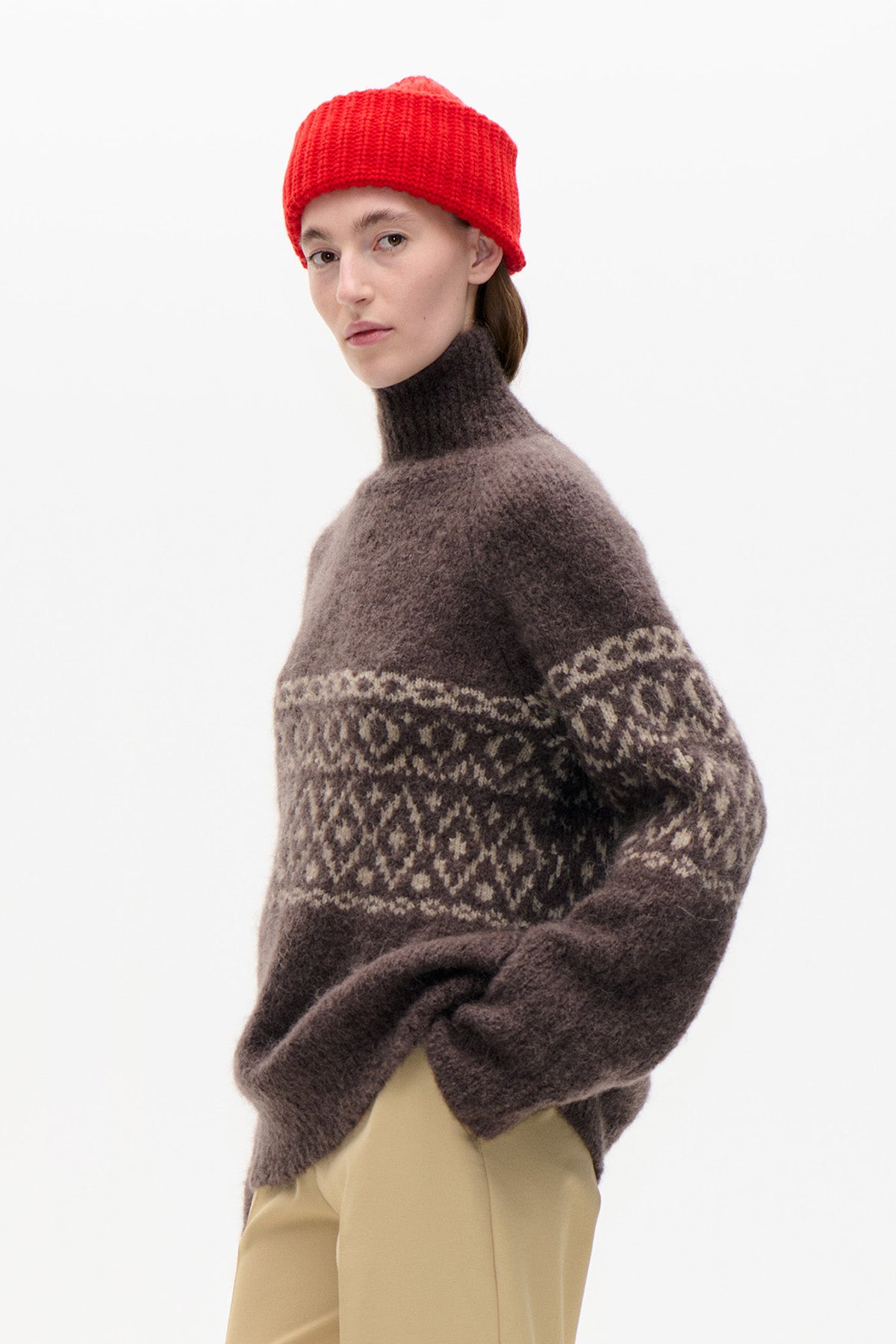 Model wearing a brown high-neck sweater with cream geometric pattern, a bright red beanie and beige trousers.