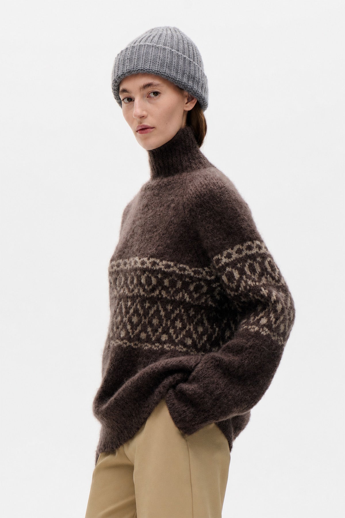 Model wearing a dark brown high-neck sweater with cream geometric patterned bands, beige trousers and a grey ribbed beanie.