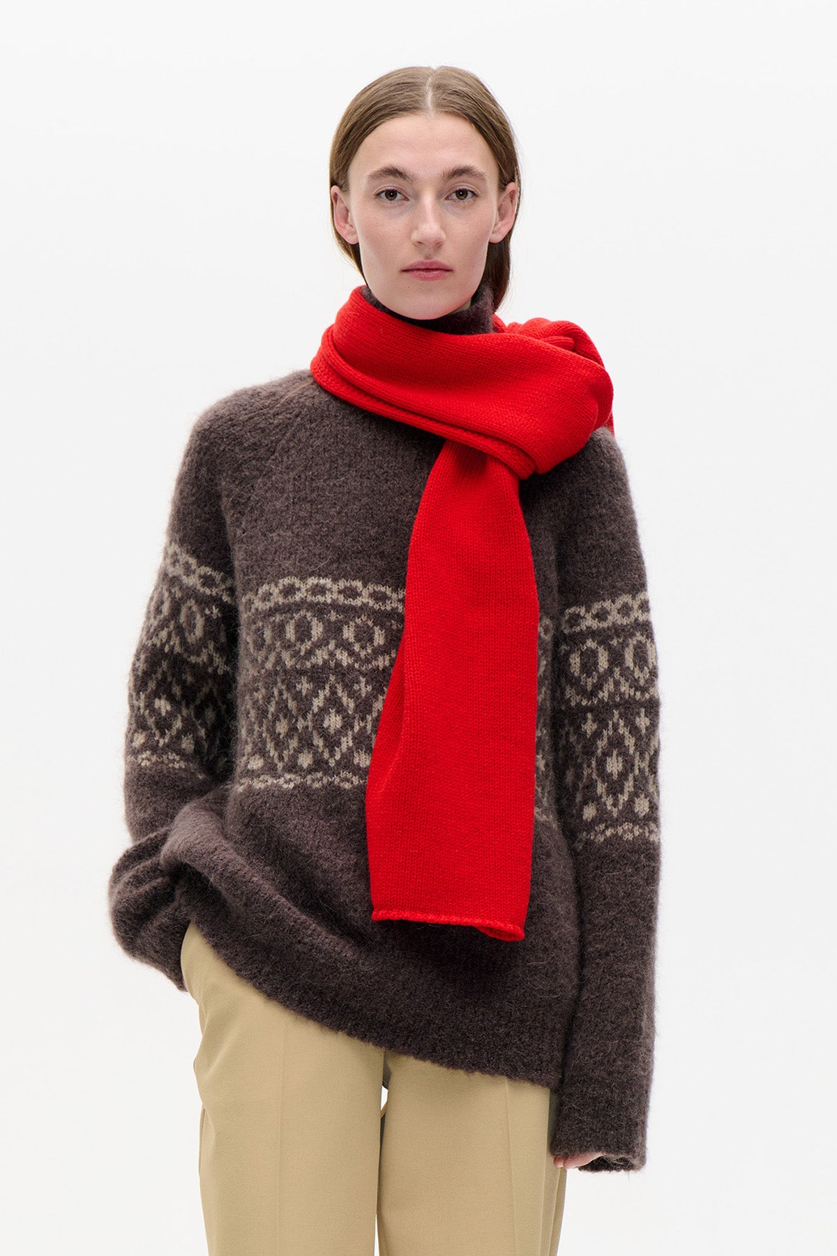Front view of a model wearing a dark brown patterned knit sweater, bright red scarf and beige trousers against a white background.