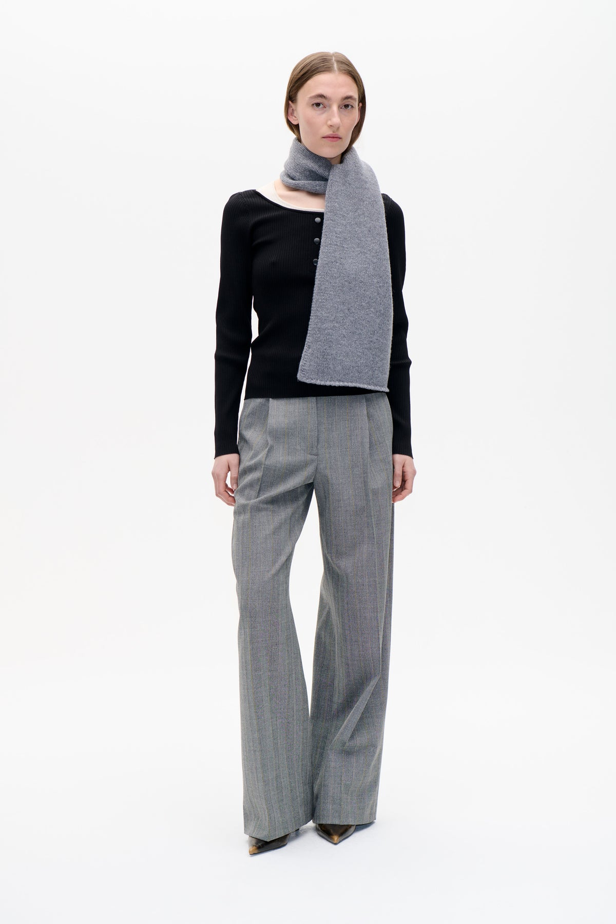 Model wearing a long heather-gray knitted scarf over a black ribbed button-front top and gray pinstriped wide-leg trousers against a white studio backdrop.