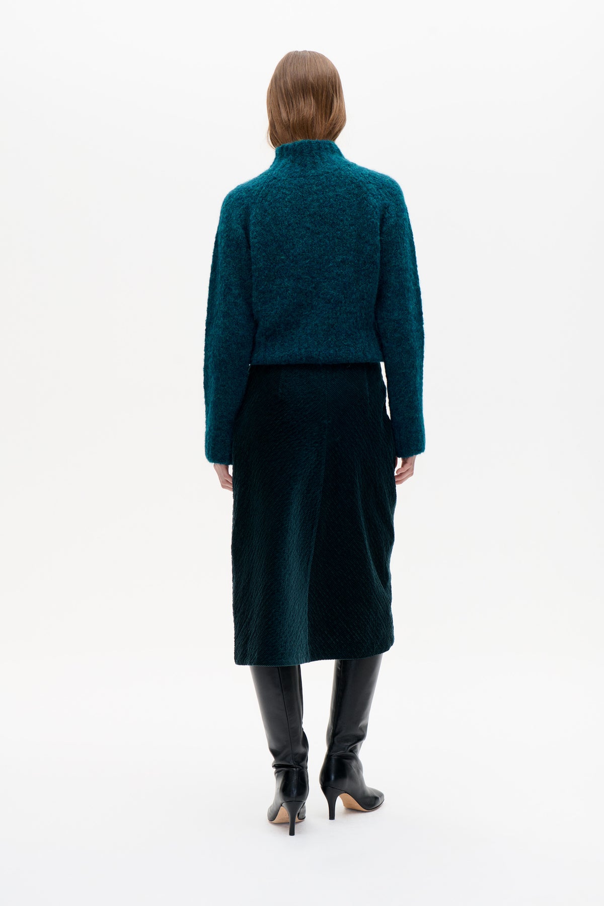 Back view of a model wearing a teal textured high-neck sweater with long sleeves and a dark skirt.