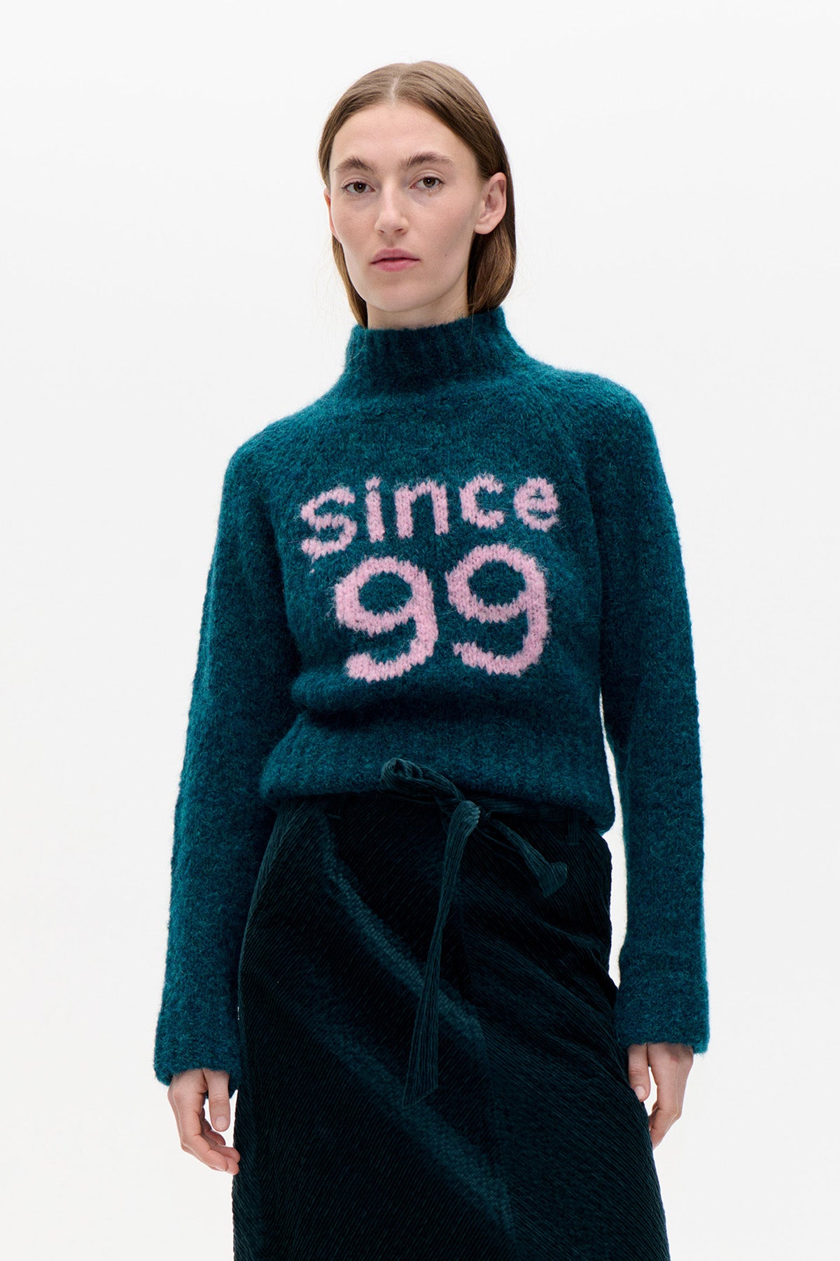 Front view of a teal high-neck sweater with pink “since 99” lettering, paired with a dark textured skirt.