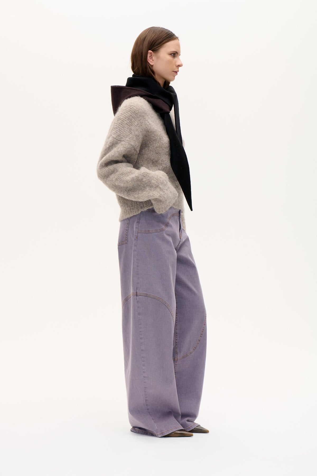 Side-profile fashion image featuring a taupe knit sweater, lavender-gray wide-leg jeans with curved contrast stitching, and a black and plum scarf.