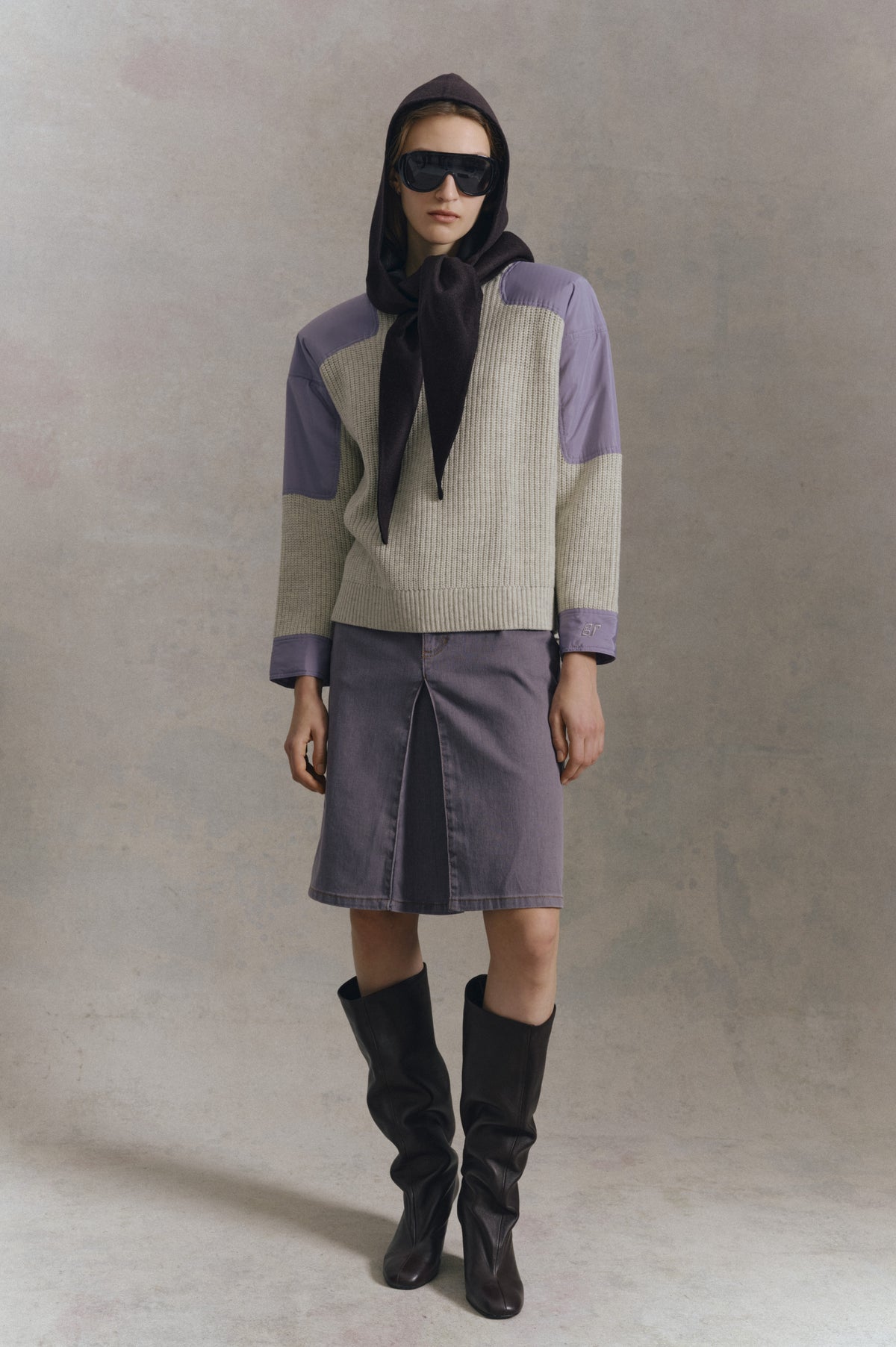Front-facing campaign portrait of a model in a cream ribbed knit vest over a lavender-sleeved top, purple denim skirt, dark scarf and tall black boots.