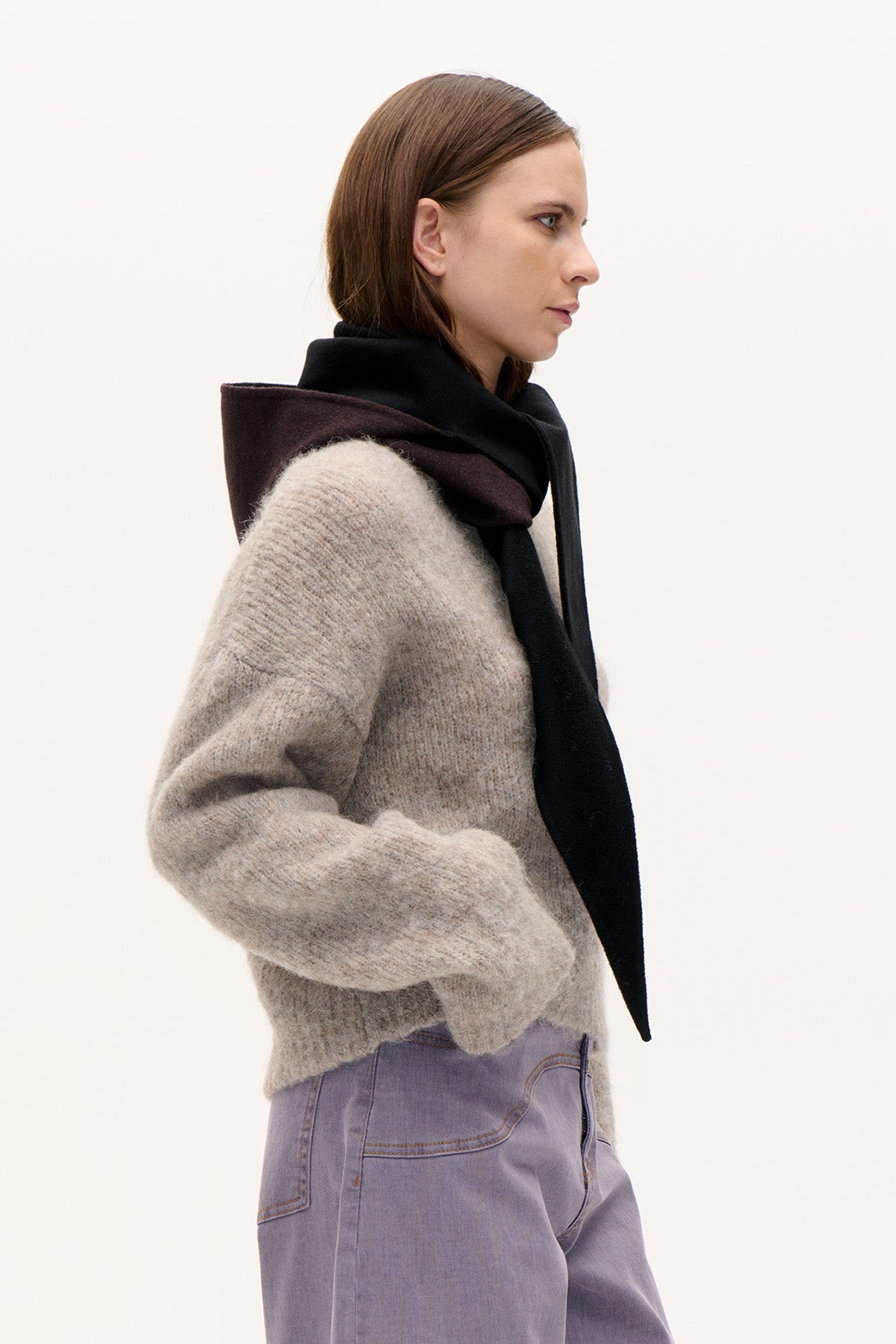 Side view of a model wearing a textured beige knit sweater, a long black scarf with a dark plum panel, and pale lilac jeans.