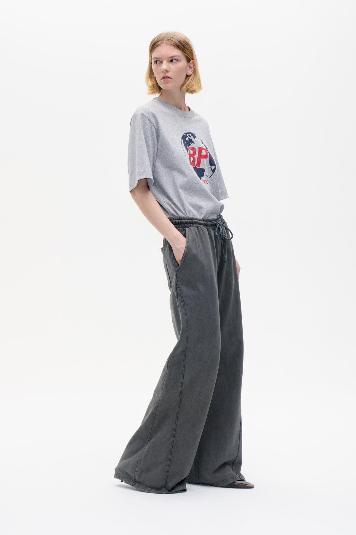 Model wearing a light grey T-shirt with a globe and red BP graphic, paired with charcoal wide-leg drawstring trousers.