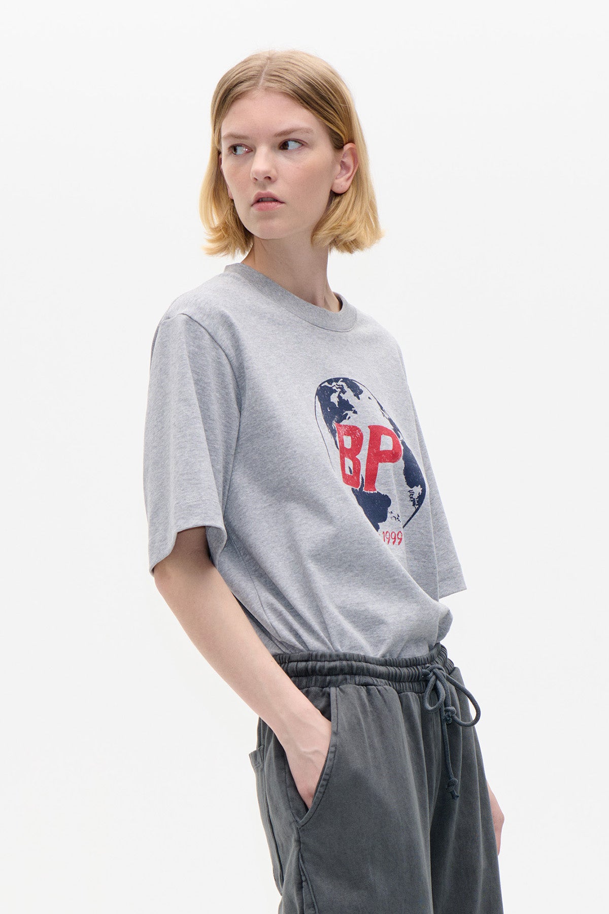 Model wearing a light grey short-sleeved T-shirt with a globe graphic and red lettering, paired with dark grey drawstring trousers.