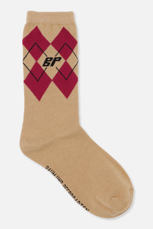 Beige crew-sok met bordeauxrode argyle-ruitjes en een klein zwart monogram, schuin afgebeeld.