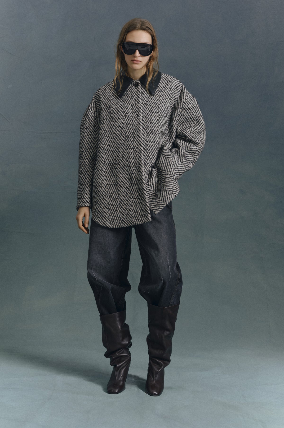 Model wearing an oversized cream and black herringbone-patterned top with a black collar, dark jeans, tall boots and sunglasses against a grey backdrop.