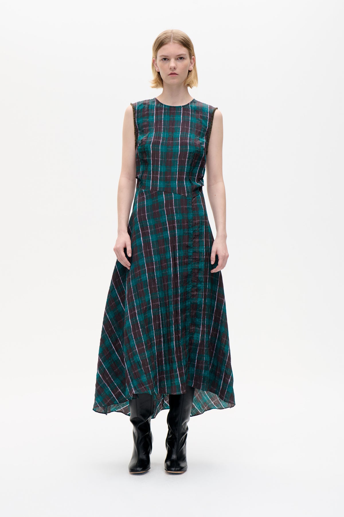 Front view of a model wearing a sleeveless teal and plum checked dress with a high-low hem and black knee-high boots.